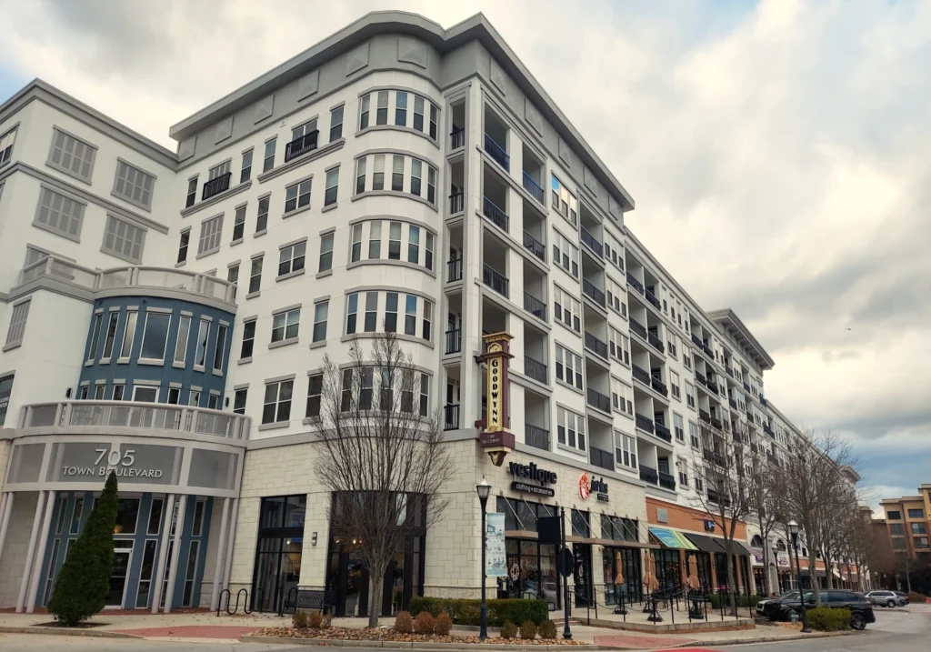 Photo showing a 6 story apartment building with various local shops on the ground level. A sidewalk runs in front of the stores and is lined with shrubs, trees, and rock landscaping.